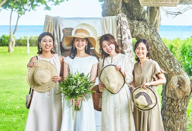 リゾートウエディングの屋外会場にて、笑顔で並ぶ4名の女性ゲスト。ベージュやオフホワイトの軽やかなワンピースを着用し、ストローハットやグリーン中心のブーケを手に持った華やかな装い。背景には青い海と豊かな緑が広がる開放的なロケーション。