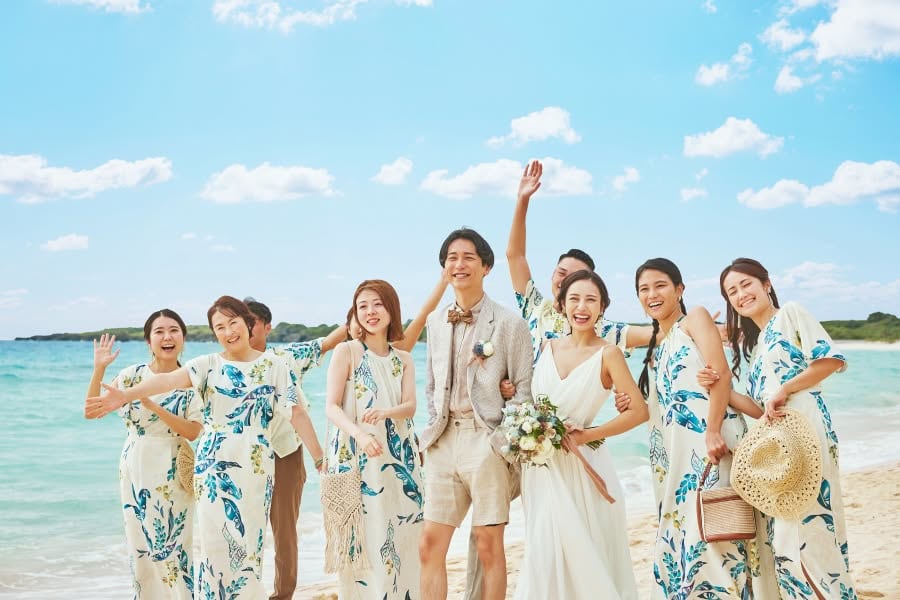 透き通る青い海と空の下、お揃いのかりゆしウェアを纏ったゲストたちに囲まれて笑顔を見せる新郎新婦のウエディングフォト。ハーフパンツスタイルの新郎と、ナチュラルな白いドレスを着た新婦が、友人や家族とリラックスした雰囲気で楽しむ、沖縄らしいビーチウエディングのワンシーンです。