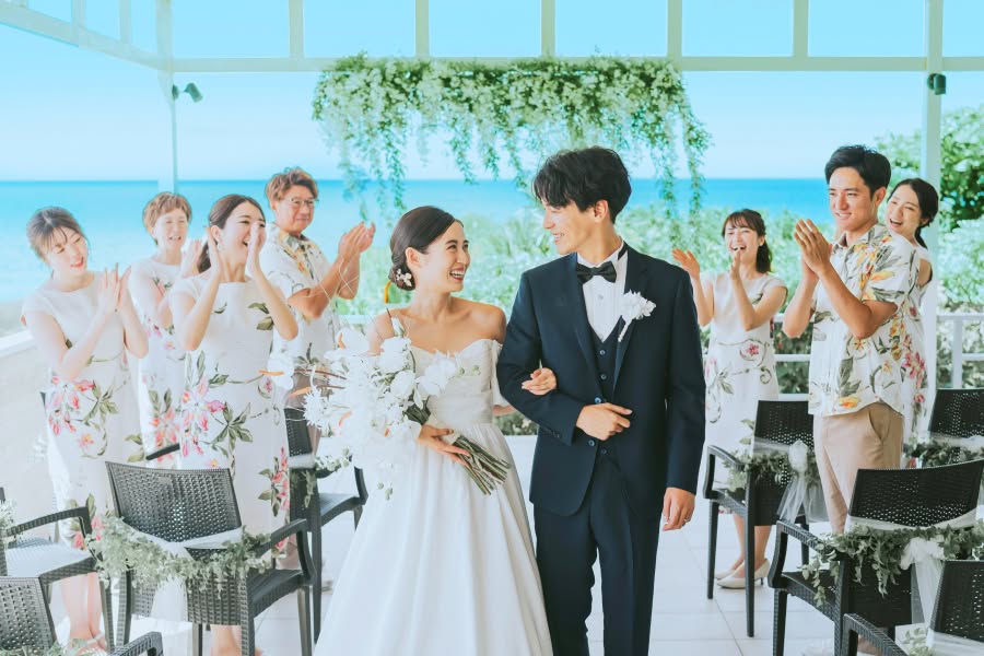 開放的なガゼボで、青い海を背景に参列者から拍手で祝福される新郎新婦のウエディングフォト。ネイビーのタキシード姿の新郎と、白いブーケを手に笑顔を見せる新婦が、かりゆしウェアを纏ったゲストに囲まれた幸せな瞬間です。