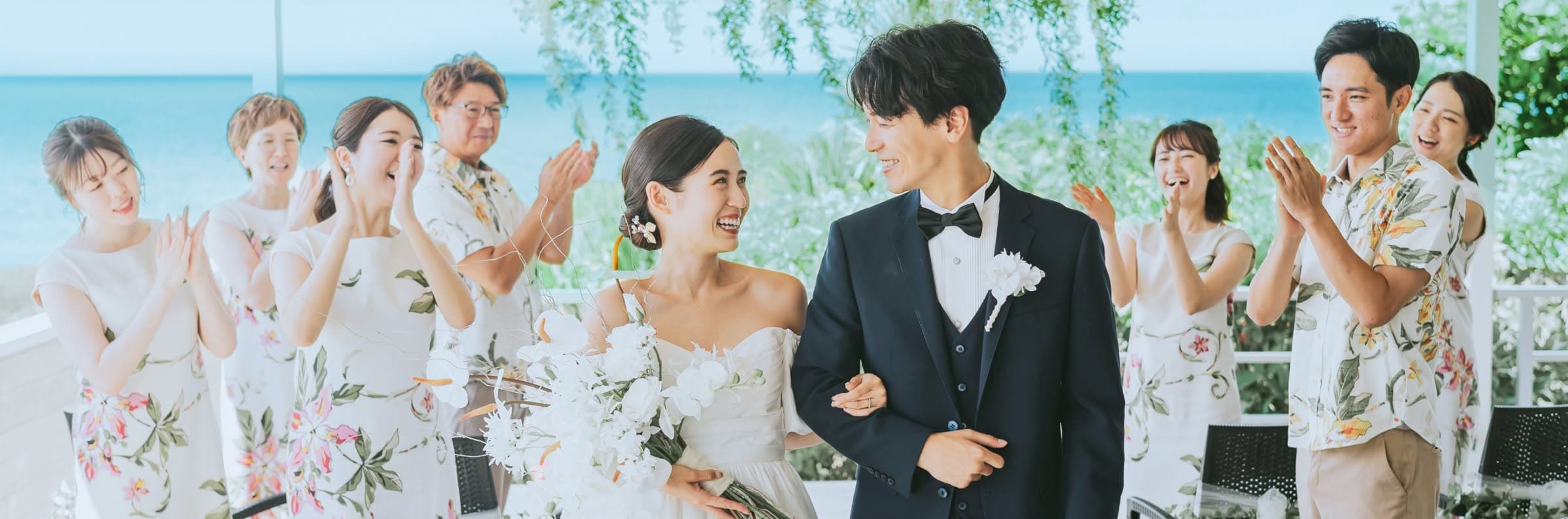 エメラルドグリーンの海を背景に、参列者からの温かい拍手に包まれて歩く新郎新婦のウエディングフォト。ネイビーのタキシード姿の新郎と、白い胡蝶蘭のブーケを抱えた笑顔の新婦が寄り添い、かりゆしウェアを纏ったゲストと共に幸せを分かち合う感動的な挙式シーンです。
