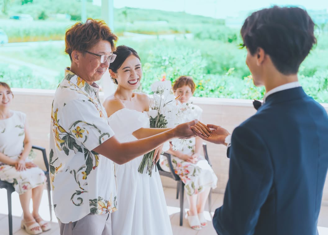 明るいチャペルで、かりゆしウェアを纏った父親（親族）に見守られながら、新郎が笑顔の新婦の手を優しく取るウエディングフォト。家族の絆と喜びが溢れる、沖縄らしい温かな挙式セレモニーの瞬間を捉えた一枚です。