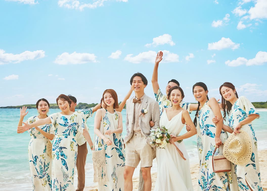 エメラルドグリーンの海が広がるビーチで、お揃いのかりゆしウェアを纏ったゲストたちと並んで笑顔を見せる新郎新婦のウエディングフォト。青い空と白い砂浜に、色鮮やかな衣装が映える、沖縄らしい開放感あふれる集合カットです。
