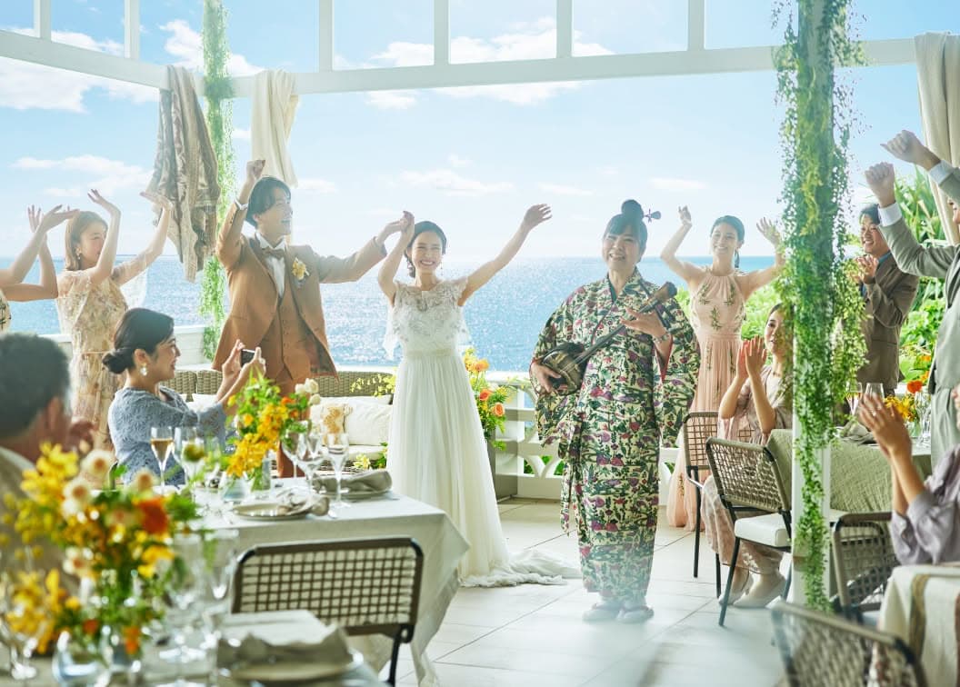 開放的なテラスで、三線の生演奏に合わせてゲストと共にカチャーシーを踊る新郎新婦のウエディングフォト。青い海を背景に、キャメル色のタキシード姿の新郎とレースが美しいドレス姿の新婦が満面の笑みで手を挙げ、沖縄らしい賑やかな祝福に包まれている挙式シーンです。
