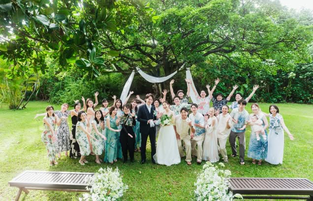 豊かな緑に囲まれた大きな樹の下で、新郎新婦を囲んでゲスト全員が両手を広げてポーズをとる賑やかな集合写真。色とりどりのかりゆしウェアやドレスを纏った参列者が、開放的なガーデンで心からの祝福を贈るアットホームなウエディングシーンです。