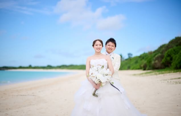 どこまでも続く白い砂浜ときらめく青い海を背景に、純白のドレスとタキシード姿で寄り添う新郎新婦のウエディングフォト。ティアラをつけた新婦が白い胡蝶蘭のブーケを手に、新郎と穏やかな笑顔を見せる、沖縄のロケーションを最大限に活かした美しいビーチ撮影の一枚です。