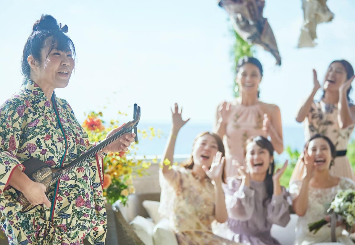 沖縄の伝統的な紅型（びんがた）衣装を纏った奏者の三線演奏に合わせ、新婦とゲストが手拍子をして盛り上がるウエディングフォト。開放感のあるテラスで、青い海を背景に沖縄らしいアットホームで賑やかな祝福を受ける挙式シーンの一枚です。