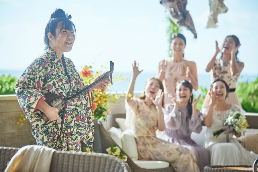 明るい陽射しが差し込む開放的なテラスで、沖縄の伝統衣装を纏った奏者の三線演奏を楽しむゲストと新婦のウエディングフォト。青い海を背景に、ゲストたちの賑やかな手拍子と笑顔に包まれながら、沖縄らしいアットホームな祝福を受ける挙式シーンの一枚です。