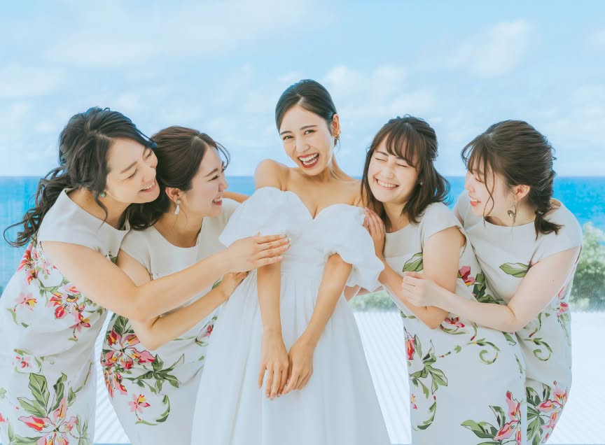 きらめく青い海を背に、お揃いのかりゆしワンピースを着た4人のブライズメイドに囲まれて満面の笑みを浮かべる新婦のウエディングフォト。オフショルダーの白いドレスを纏った新婦を友人たちが優しく祝福する、仲睦まじく華やかなリゾート感溢れる一枚です。