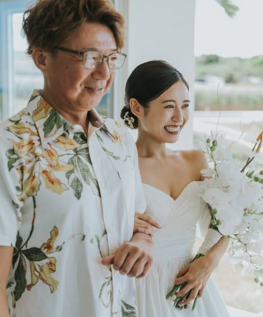 自然光が差し込むチャペル内で、かりゆしウェアを着用した父親と腕を組み、満面の笑みを浮かべてバージンロードを歩む新婦のウエディングフォト。白い胡蝶蘭のブーケを手に、幸せ溢れる表情で一歩を踏み出す感動的な挙式シーンの一枚です。