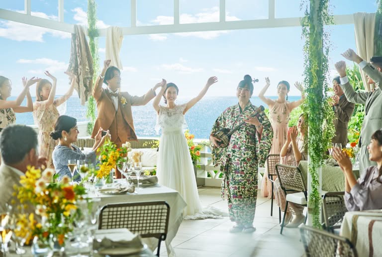 開放的なテラスで、三線の生演奏に合わせてゲストと共にカチャーシーを踊る新郎新婦のウエディングフォト。青い海を背景に、キャメル色のタキシード姿の新郎とレースが美しいドレス姿の新婦が満面の笑みで手を挙げ、沖縄らしい賑やかな祝福に包まれている挙式シーンです。