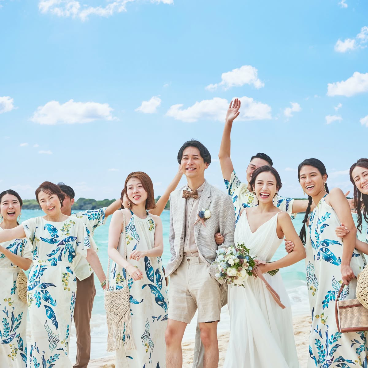 透き通る青い海と空の下、お揃いのかりゆしウェアを纏ったゲストたちに囲まれて笑顔を見せる新郎新婦のウエディングフォト。ハーフパンツスタイルの新郎と、ナチュラルな白いドレスを着た新婦が、友人や家族とリラックスした雰囲気で楽しむ、沖縄らしいビーチウエディングのワンシーンです。