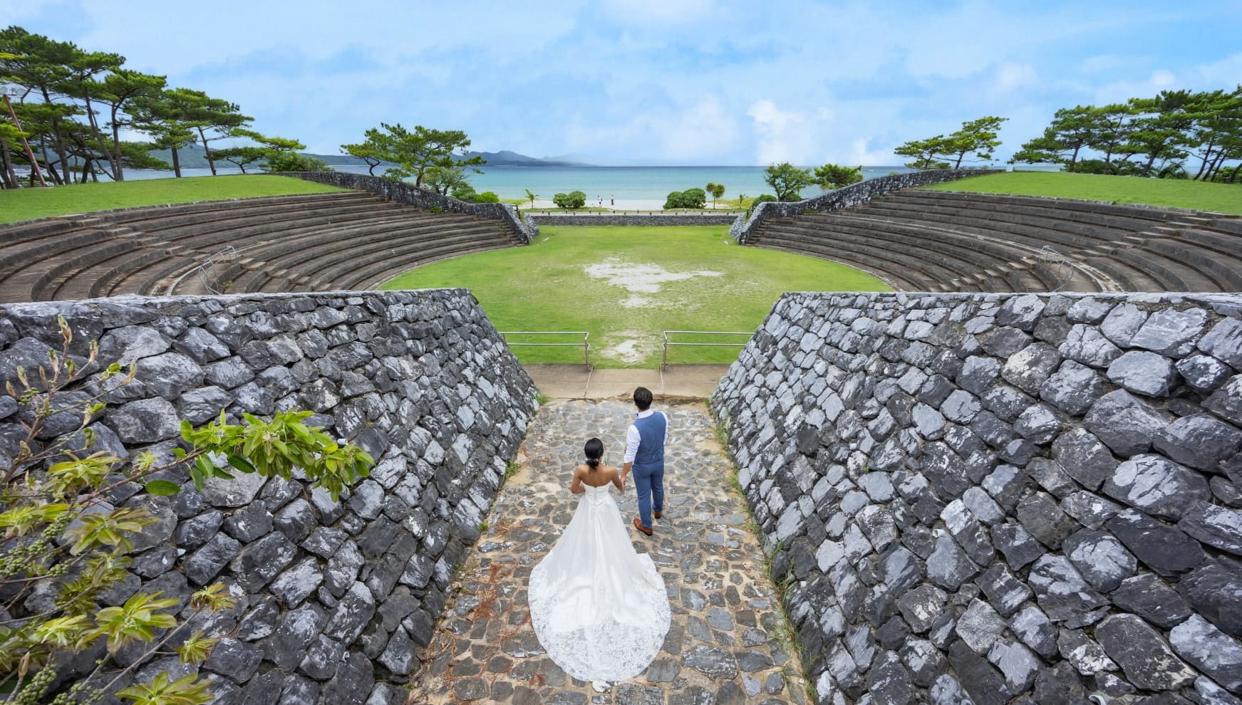 壮大な石畳の壁がそびえるロケーションで、手を繋ぎ海を眺めている新郎新婦