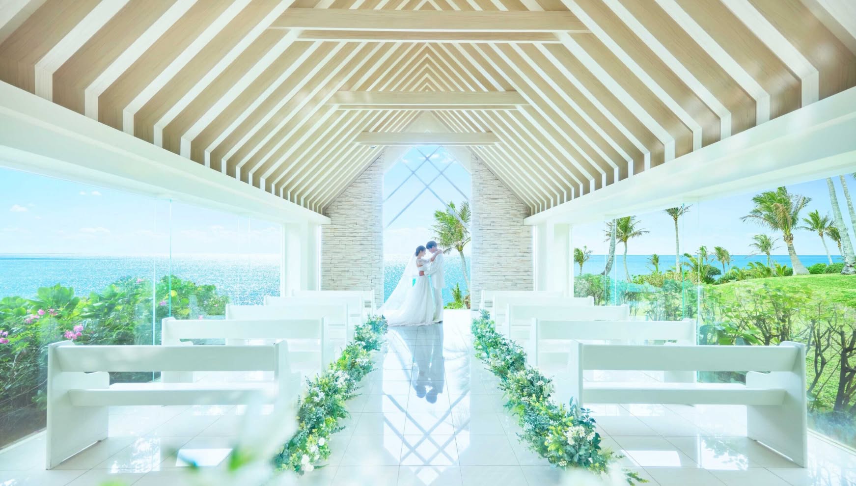 沖縄のムーンビーチにある、自然光が降り注ぐ真っ白なチャペル。正面に青い海と空が広がる開放的な空間で、ウエディングドレスとタキシード姿の新郎新婦が祭壇前で見つめ合う、神聖で幻想的な結婚式の風景です。
