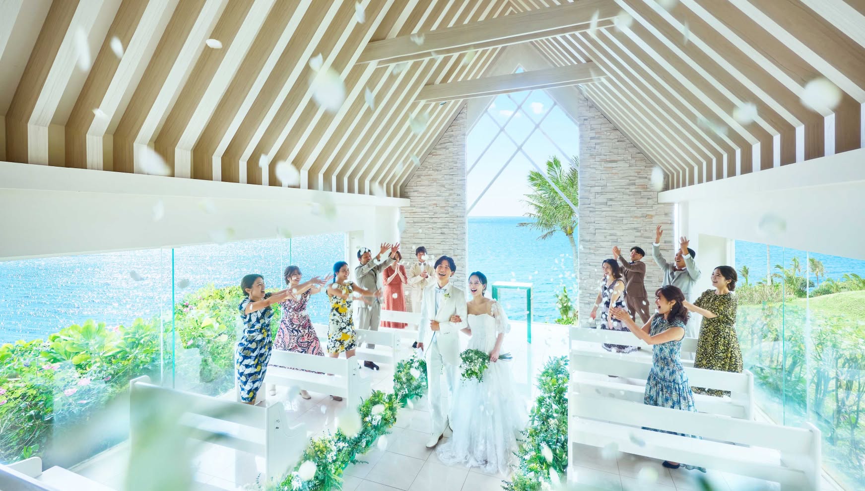 沖縄のムーンビーチにある、海を一望する白亜のチャペル。挙式を終えたタキシードとウエディングドレス姿の新郎新婦が笑顔でバージンロードを歩き、参列したゲストから祝福の花吹雪（フラワーシャワー）を受ける幸せな瞬間です。