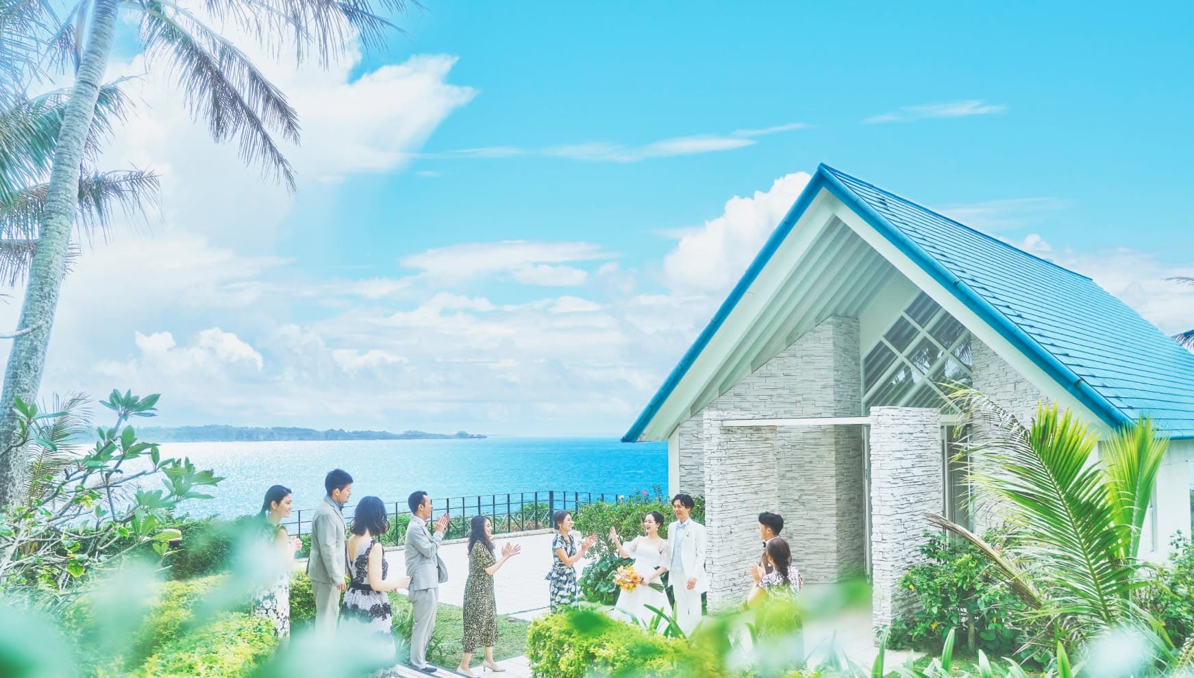 沖縄の海を一望する高台に立つ、青い屋根のモダンなチャペル外観。挙式を終えたばかりのウエディングドレス姿の新婦とタキシード姿の新郎が、青い空と海を背景に、ゲストから祝福の拍手を受けている幸せな光景です。