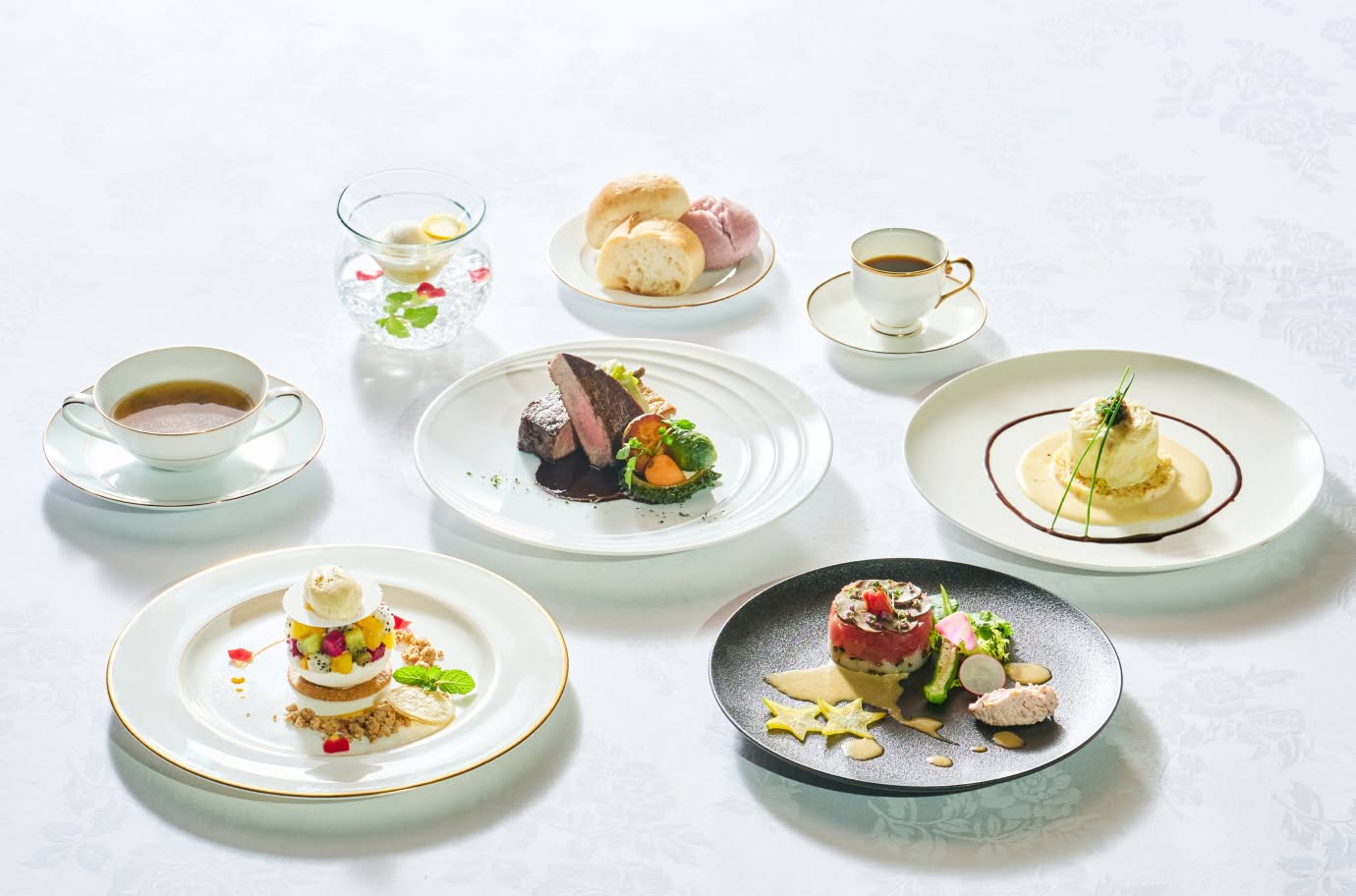 白いテーブルクロスの上のウエディングフルコース料理。牛フィレ肉のステーキ、鮮やかな前菜、デザート、スープ、パン、コーヒーが美しく並んでいます。沖縄の食材を活かし、色彩豊かに盛り付けられた婚礼料理の全景です。