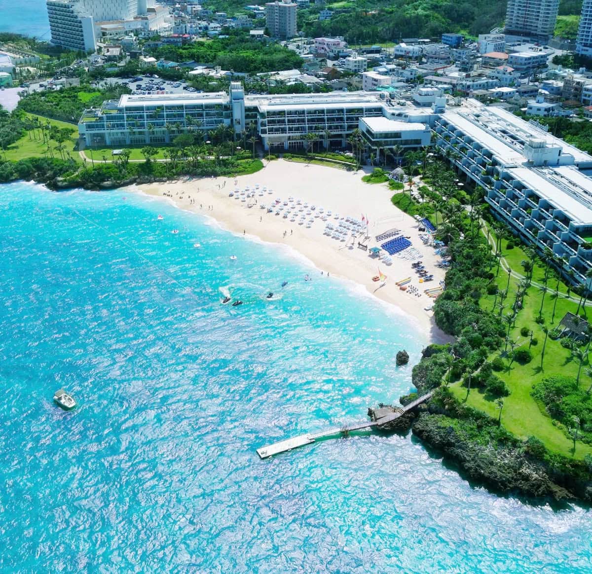 沖縄の本島にあるホテルムーンビーチの全景。エメラルドグリーンの美しい海に囲まれた三日月形の白い砂浜と、緑豊かな敷地に建つリゾートホテルの建物を上空から捉えた、開放感あふれるウエディングロケーションの俯瞰。