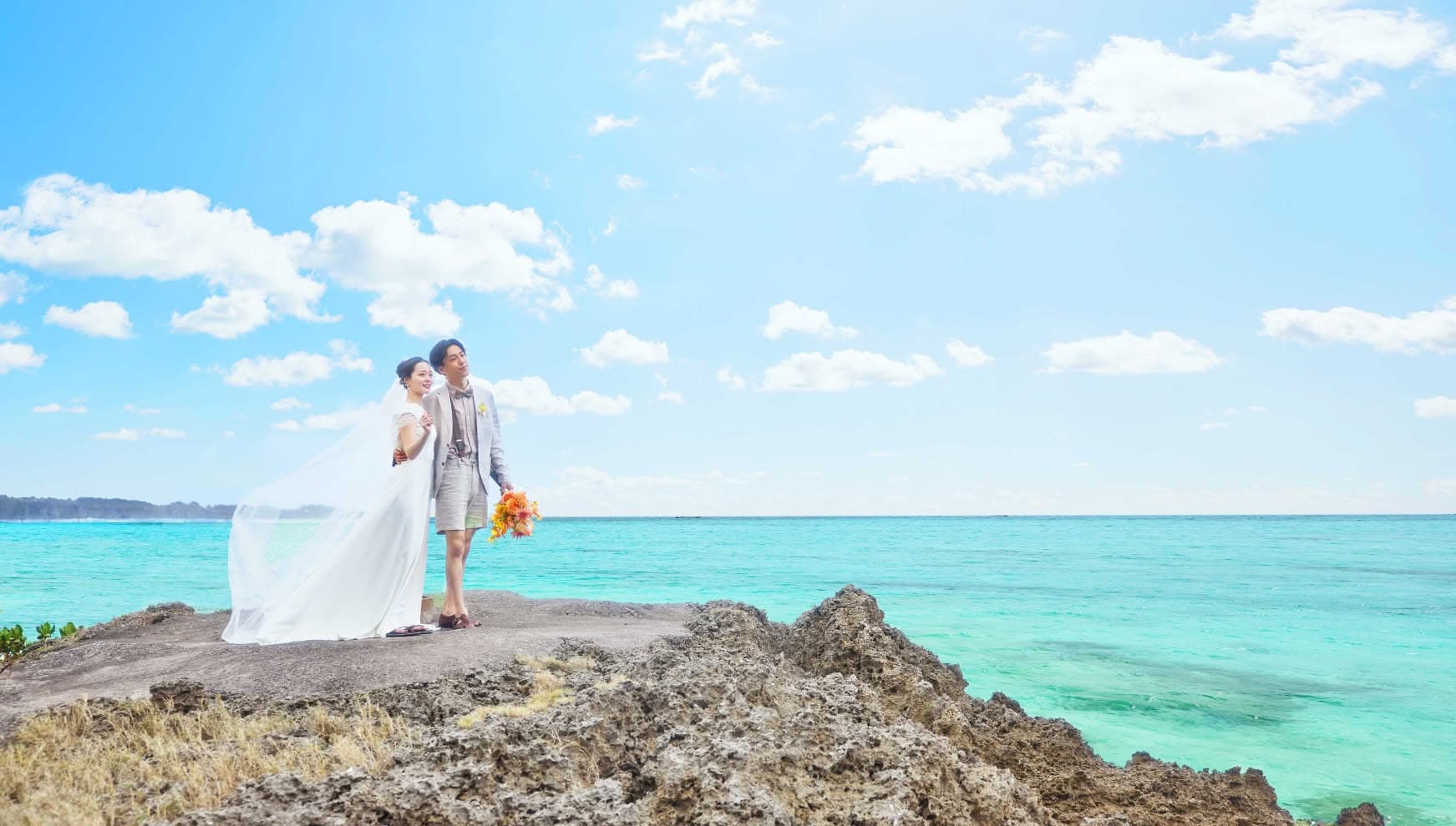沖縄の鮮やかなエメラルドグリーンの海を背景に、ゴツゴツとした岩場に立つ新郎新婦。風に吹かれてなびく長いベールが美しいウエディングドレス姿の新婦と、ハーフパンツのカジュアルなタキシード姿の新郎が空を見上げるロケーションフォト。