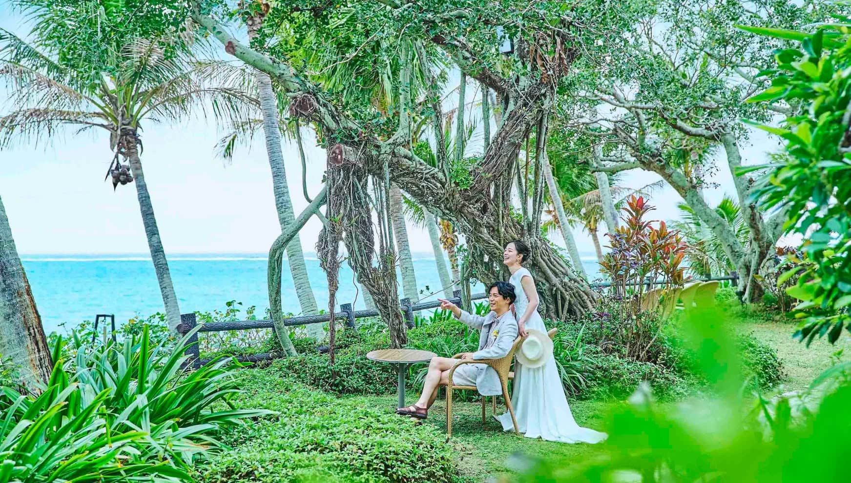 沖縄のムーンビーチにある緑豊かなガーデンで、海を背景にリラックスする新郎新婦。ガジュマルの木の下で、椅子に座るカジュアルなタキシード姿の新郎と、その横で笑顔を見せるウエディングドレス姿の新婦のロケーションフォト。