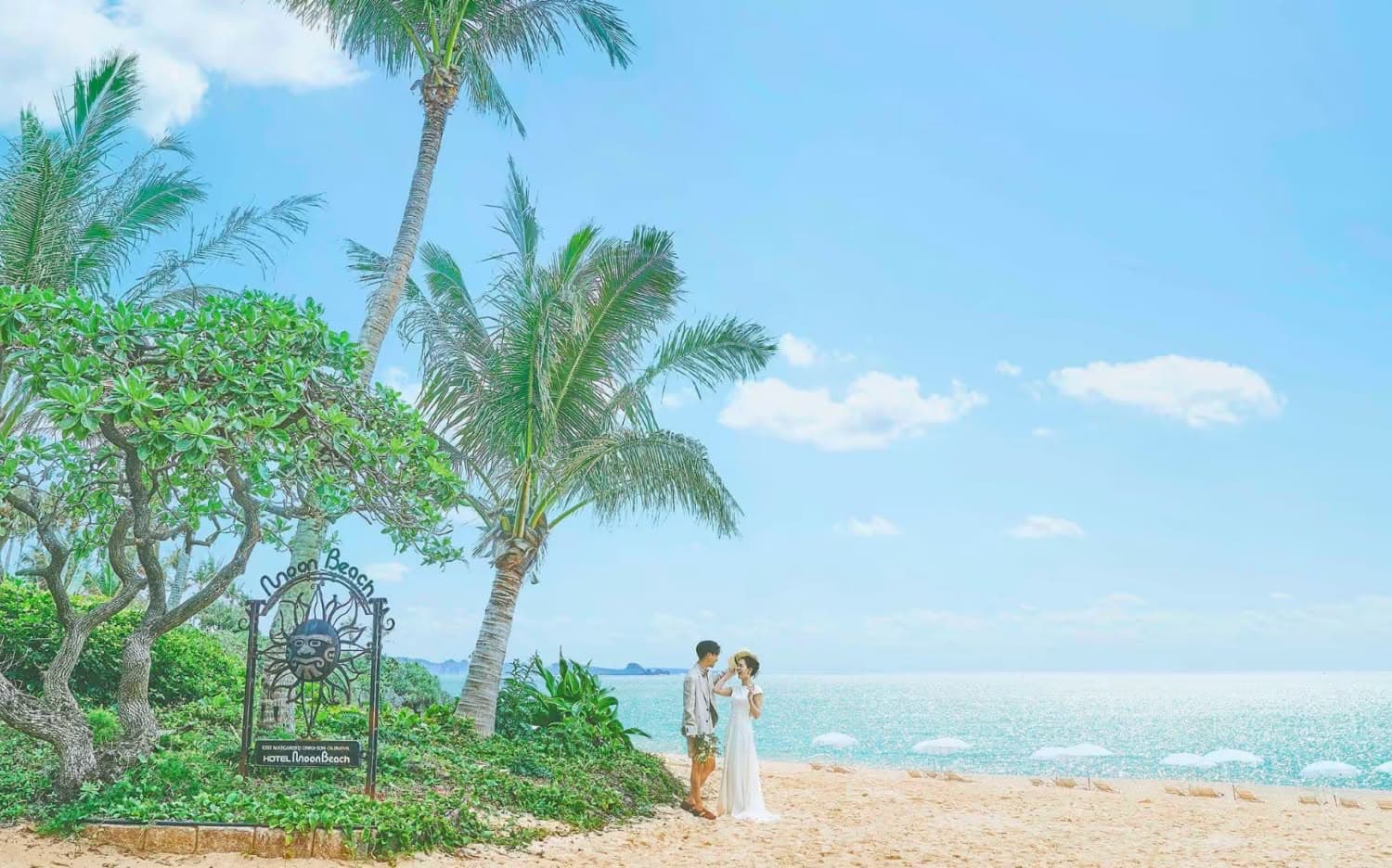 沖縄の海を望む開放的なテラスでのウエディングパーティー。白いタキシードとウエディングドレス姿の新郎新婦が笑顔で入場し、ゲストが拍手で祝福する様子。背景には青い海とヤシの木が広がり、南国らしい披露宴の情景です。