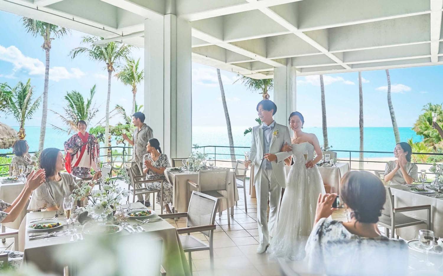 沖縄の海を望む開放的なテラスでのウエディングパーティー。白いタキシードとウエディングドレス姿の新郎新婦が笑顔で入場し、ゲストが拍手で祝福する様子。背景には青い海とヤシの木が広がり、南国らしい披露宴の情景です。