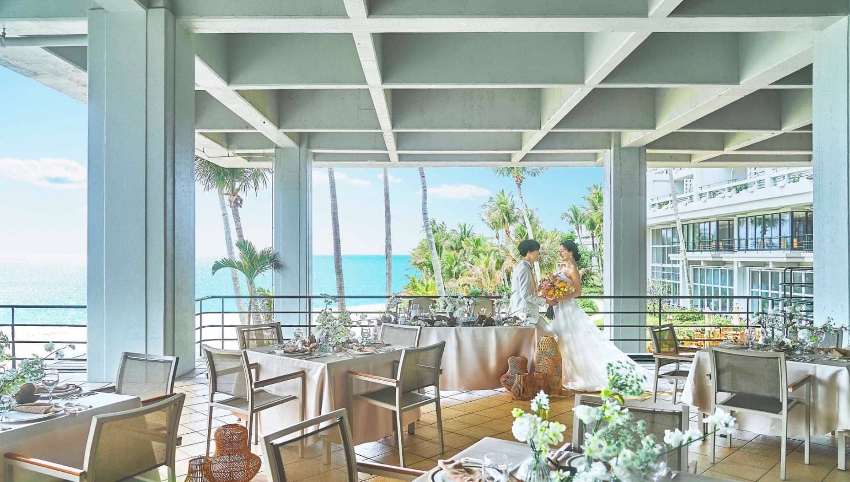 沖縄の海と椰子の木を望む開放的なテラスでの披露宴会場。白いテーブルクロスと装花が整えられた席の奥で、ウエディングドレスとタキシード姿の新郎新婦が寄り添う様子。リゾートの風を感じる明るいウエディングシーンです。