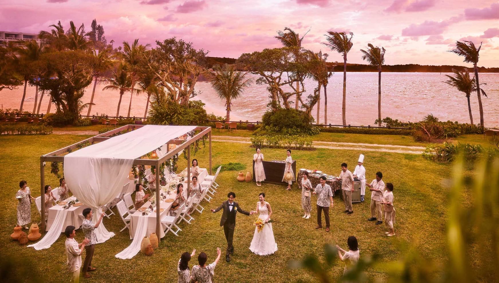 夕暮れ時の広大なガーデンで行われるウエディングパーティー。夕日に染まる空と海を背景に、ウエディングドレスとタキシード姿の新郎新婦が笑顔で歩き、ゲストが温かく拍手で迎える、アットホームで開放的な披露宴の様子です。