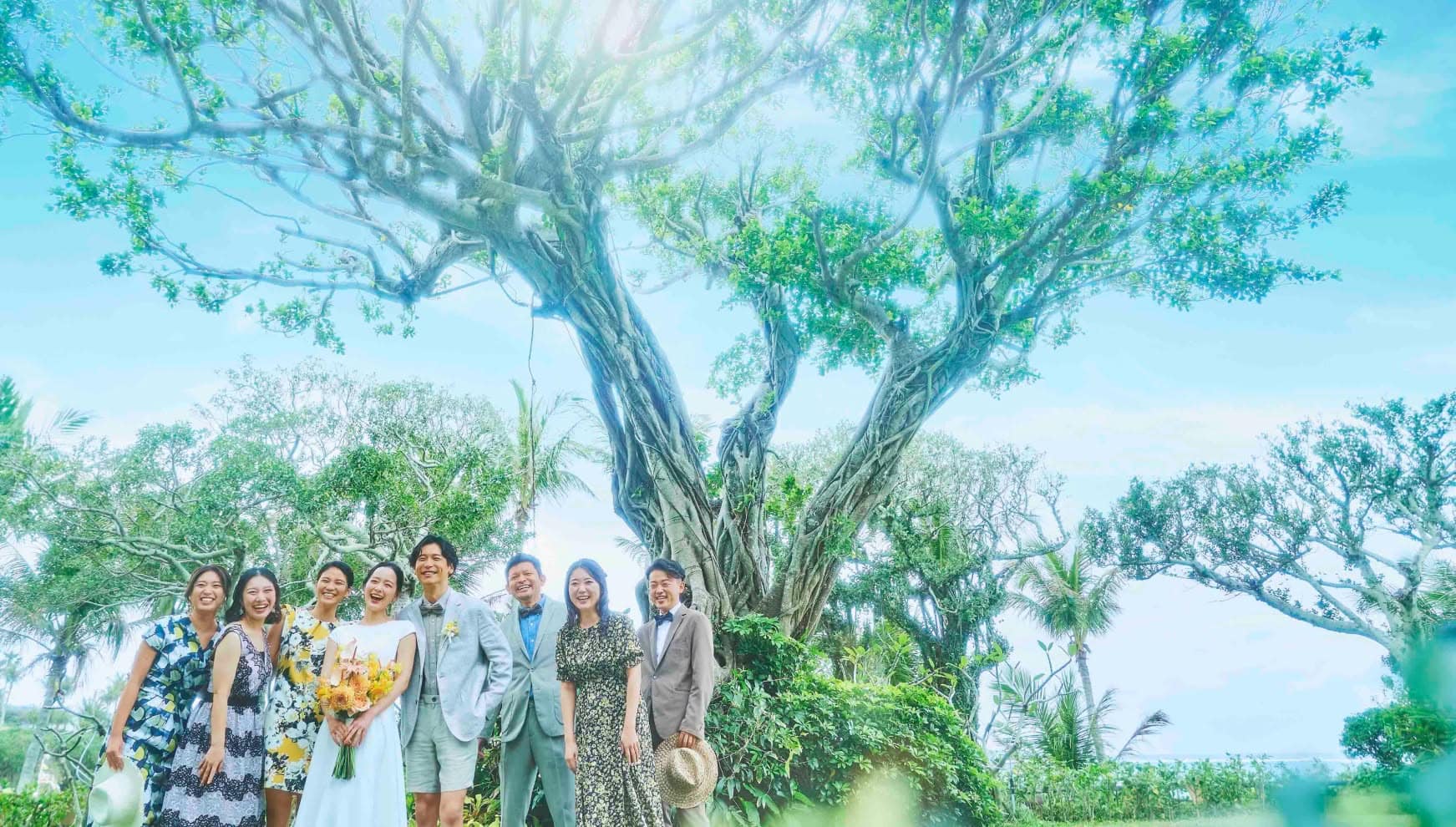 沖縄のムーンビーチにある、生命力溢れる巨大なガジュマルの木の下で、ウエディングドレス姿の新婦とカジュアルな装いの新郎、そして笑顔のゲストたちが並んで記念撮影をする、アットホームで自然豊かな集合写真。