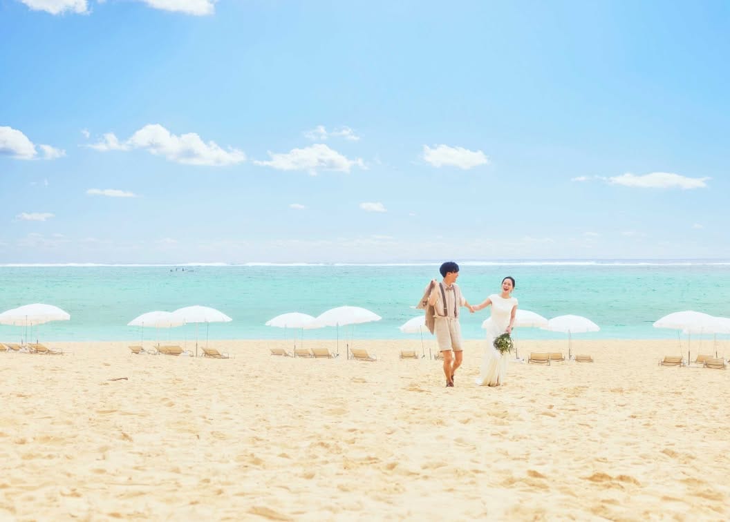 沖縄の真っ白な砂浜が広がるムーンビーチで、エメラルドグリーンの海を背に手をつないで歩く新郎新婦。ウエディングドレス姿の花嫁と、サスペンダーにハーフパンツのカジュアルな装いの新郎が笑顔で語らう、開放感あふれるビーチフォト。