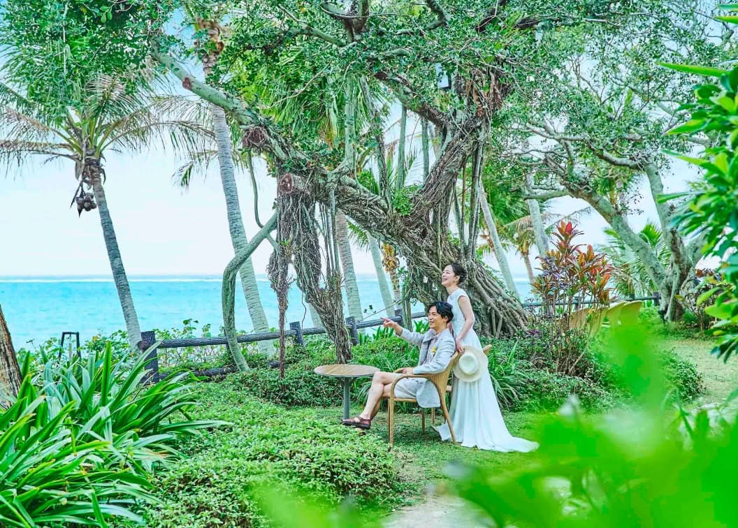 沖縄のムーンビーチにある緑豊かなガーデンで、海を背景にリラックスする新郎新婦。ガジュマルの木の下で、椅子に座るカジュアルなタキシード姿の新郎と、その横で笑顔を見せるウエディングドレス姿の新婦のロケーションフォト。