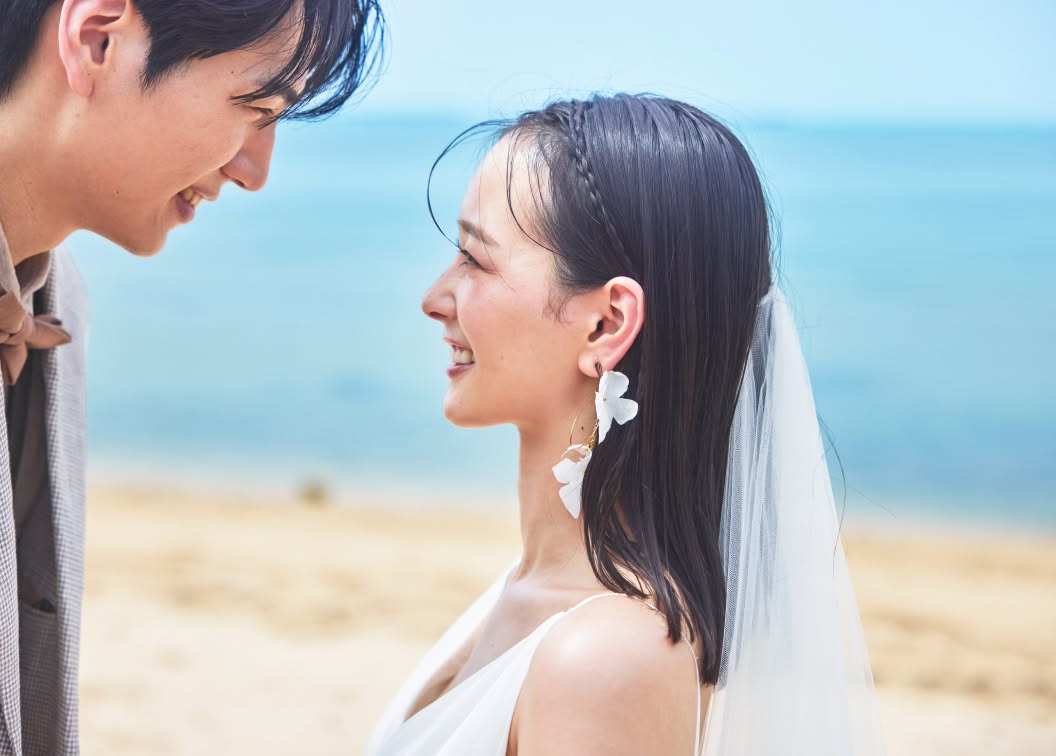 沖縄のビーチを背景に、見つめ合う新郎新婦のアップショット。白い花びらの大ぶりなピアスを身につけ、ベールを纏った笑顔の花嫁と、優しく微笑む新郎の横顔が写っています。ふたりの幸せな表情にフォーカスした、情緒的なウエディングカットです。