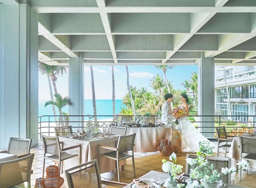 沖縄の海と椰子の木を望む開放的なテラスでの披露宴会場。白いテーブルクロスと装花が整えられた席の奥で、ウエディングドレスとタキシード姿の新郎新婦が寄り添う様子。リゾートの風を感じる明るいウエディングシーンです。