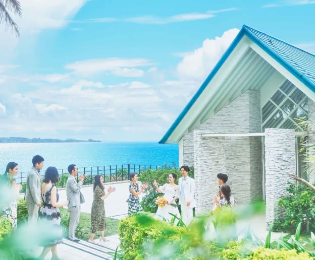沖縄の海を一望する高台に立つ、青い屋根のモダンなチャペル外観。挙式を終えたばかりのウエディングドレス姿の新婦とタキシード姿の新郎が、青い空と海を背景に、ゲストから祝福の拍手を受けている幸せな光景です。