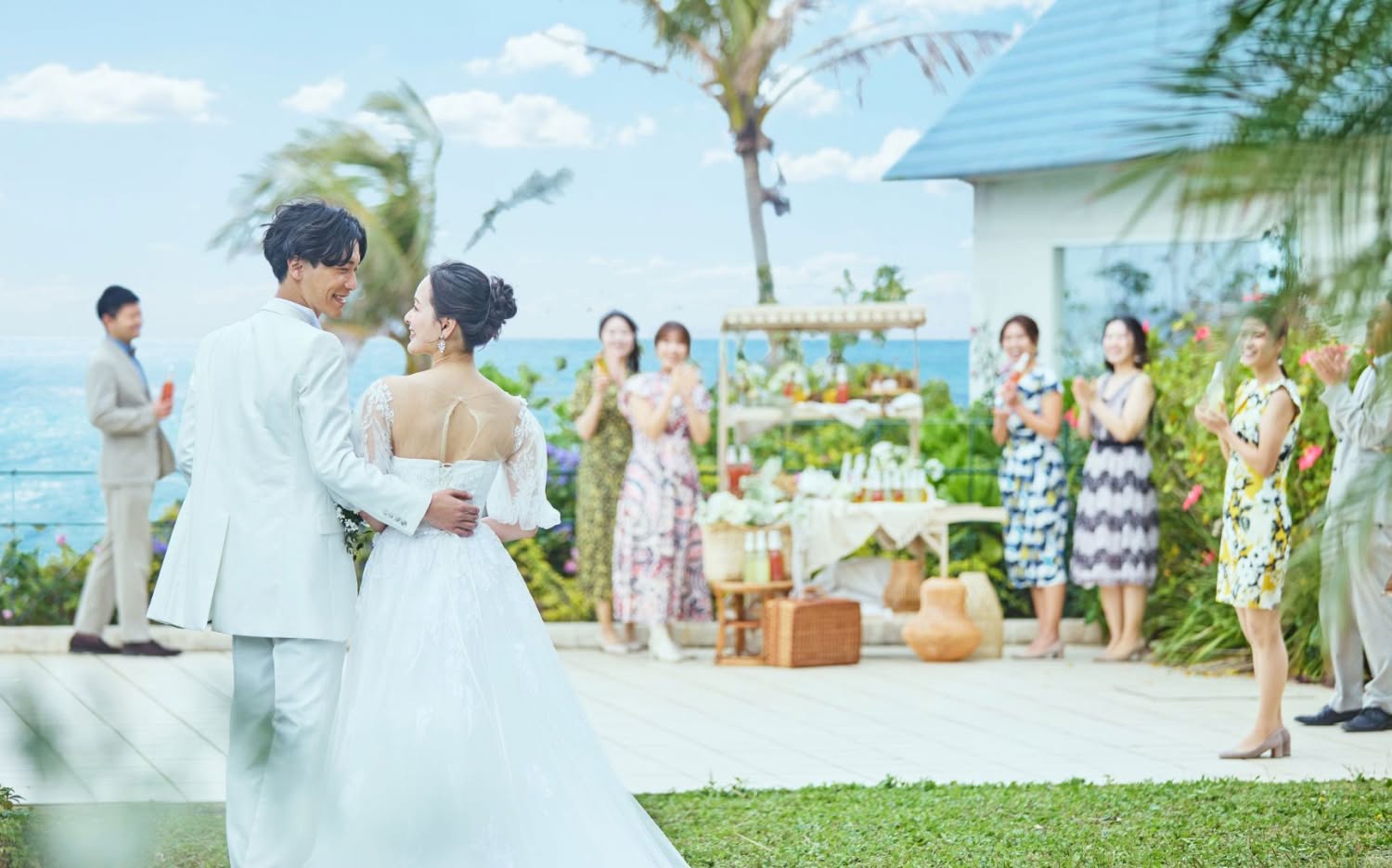 沖縄の海を一望できる開放的なテラスでのウエディングパーティー。白いタキシードとウエディングドレス姿の新郎新婦が笑顔で入場し、ゲストが温かい拍手で迎える様子。テーブルには料理や装花が並び、リゾート感溢れる披露宴の光景です。