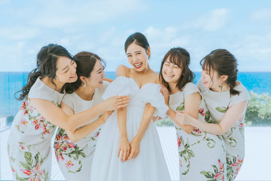 澄み渡る青い海を背景に、オフショルダーの白いウエディングドレスを纏った新婦が、お揃いのかりゆしウェアを着た4名の友人に囲まれて満面の笑みを浮かべる様子。沖縄の爽やかな風を感じさせる、ブライズメイドとの仲睦まじい記念写真。