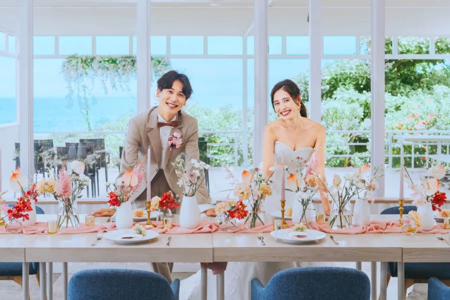 明るい光が差し込むオーシャンビューの会場で、ピンクやオレンジの装花が飾られたテーブル越しに笑顔を見せる新郎新婦。ベージュのタキシードとビスチェタイプのウエディングドレス姿の二人が、沖縄の海を背景にゲストを迎えるパーティーシーン。