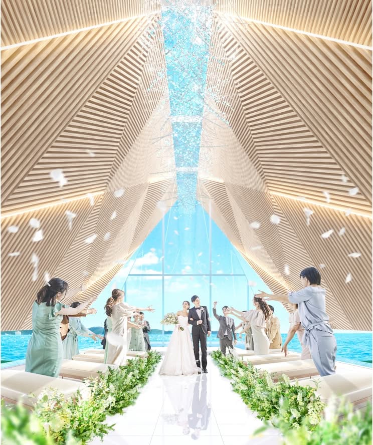 木の温もりを感じる三角形のチャペルで、青い海と空を背景にフラワーシャワーで祝福される新郎新婦。タキシードとウエディングドレス姿の二人が、ゲストに囲まれながら光り輝くバージンロードを歩む、幻想的な沖縄ウエディングの挙式シーン。