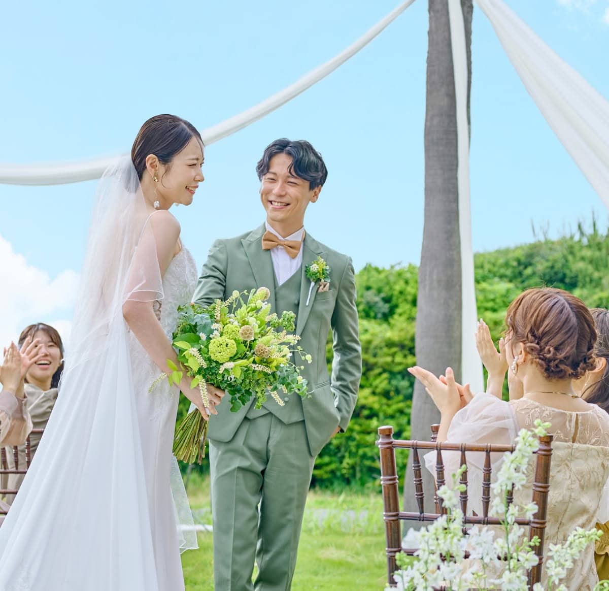 青空の下、グリーンのタキシードを着た新郎とウエディングドレス姿の新婦が、ゲストの拍手に包まれながら笑顔で見つめ合う屋外挙式の様子。沖縄の自然豊かなガーデンで、色鮮やかなブーケを手にした幸せ溢れるワンシーン。