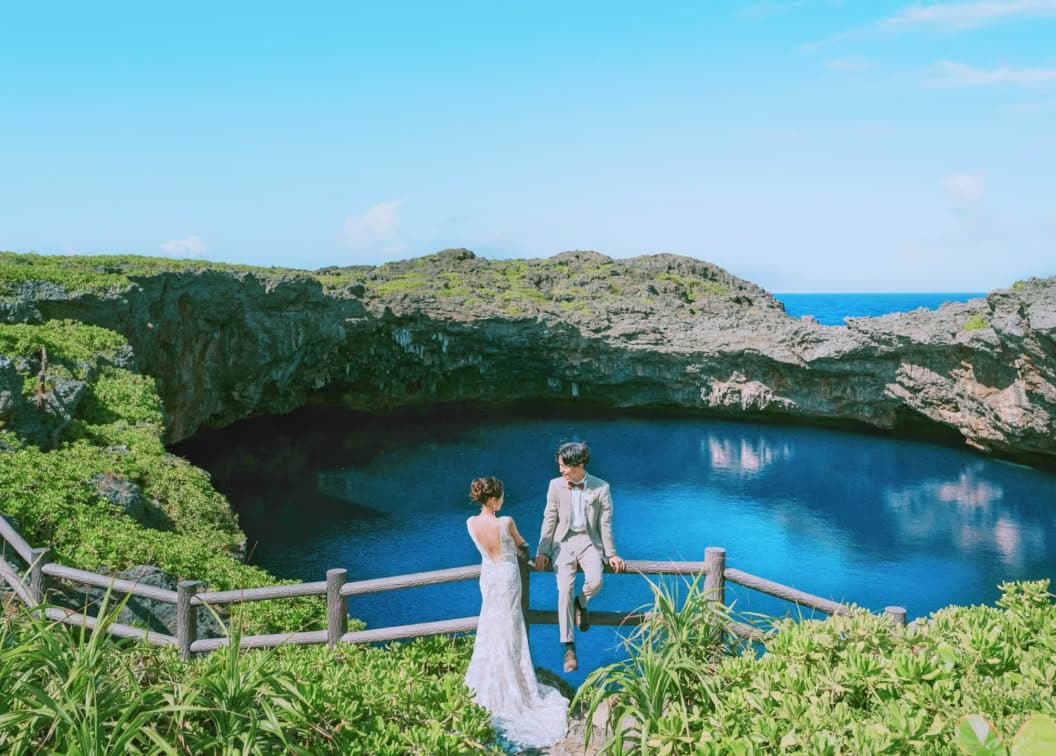 沖縄のダイナミックな断崖絶壁に囲まれた青い入り江をバックに、柵に寄り添う新郎新婦。ベージュのタキシード姿の新郎と、背中が大きく開いたマーメイドラインのウエディングドレスを纏った新婦が、大自然の中で静かに見つめ合うロケーションフォト。