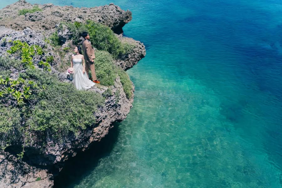 エメラルドグリーンの澄み渡る沖縄の海を背景に、切り立った崖の上に立つ新郎新婦。ブラウンのタキシードと白いウエディングドレス姿の二人が、広大な自然の中で寄り添う幻想的なロケーションフォトのワンシーン。