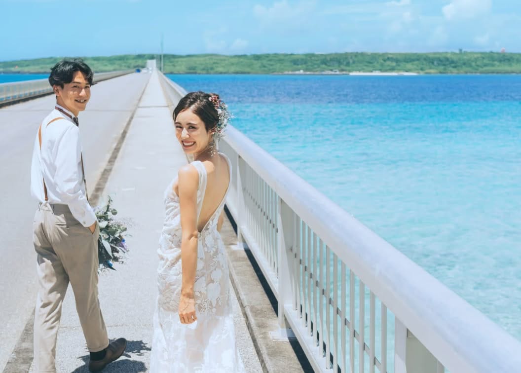 どこまでも続く青い海と空に囲まれた伊良部大橋を背景に、笑顔で振り返る新郎新婦。サスペンダー姿のカジュアルな新郎と、背中が大きく開いた刺繍入りのウエディングドレスを纏った新婦が、宮古島の絶景の中で撮影を楽しむロケーションフォト。