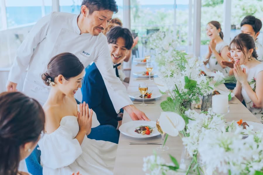沖縄の海が見える披露宴会場で、シェフから料理をサーブされる新婦の様子。オフショルダーのウエディングドレスを着た新婦とタキシード姿の新郎が、ゲストに囲まれながら笑顔で食事を楽しむ、おもてなしの心が伝わるパーティーシーン。