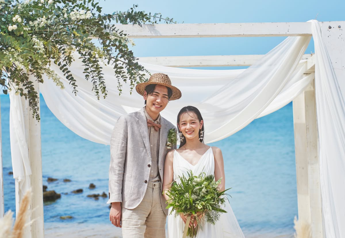 沖縄の青い海をバックに、白い布とグリーンの装花で飾られたビーチガゼボの前で微笑む新郎新婦。麦わら帽子にリネンジャケットを合わせたカジュアルな新郎と、ナチュラルなウエディングドレス姿の新婦が、南国の開放感あふれる挙式シーンを演出するウエディングフォト。