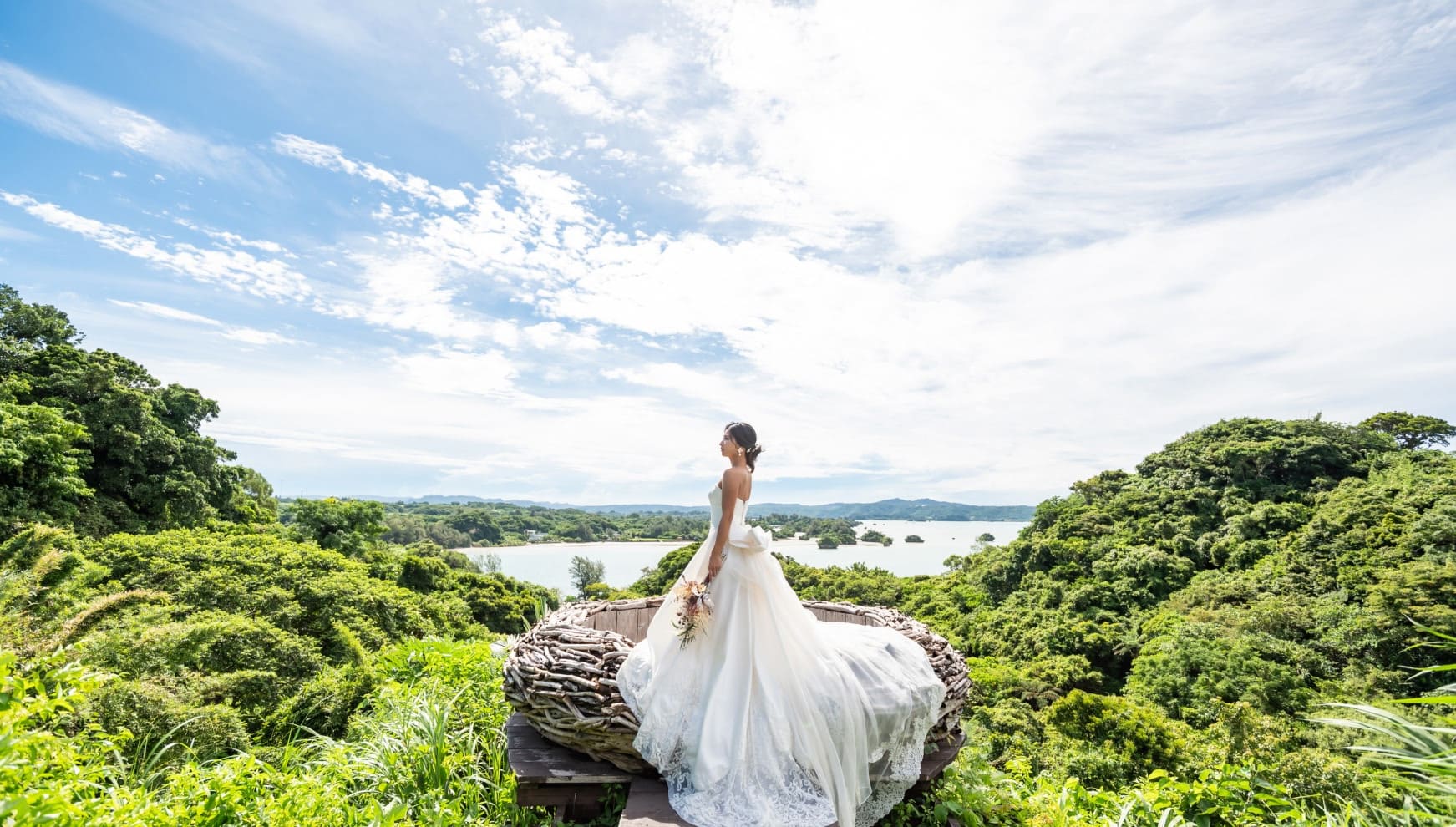 沖縄・今帰仁村の絶景ロケーション「CASA SOL（カーサ・ソル）」でのフォトウエディング。緑豊かな森と静かな海を見渡す高台にて、鳥の巣をイメージしたダイナミックな木製オブジェに立つ新婦。繊細なレースが美しいロングトレーンのドレスが風に舞う、神秘的な一枚。