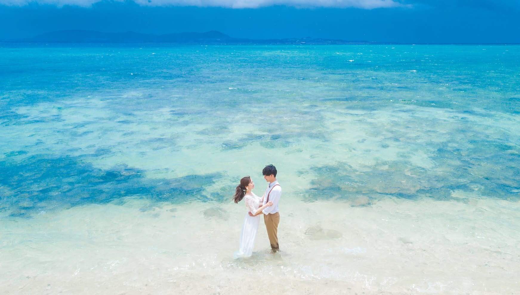 石垣島の「電信屋ビーチ」にて、透明度抜群のクリスタルブルーの海に足元を浸しながら見つめ合う新郎新婦。波打ち際の透き通るような白砂と、幾重にも重なる青のグラデーションが美しい海が広がる、幻想的で清涼感あふれるウエディングフォト。
