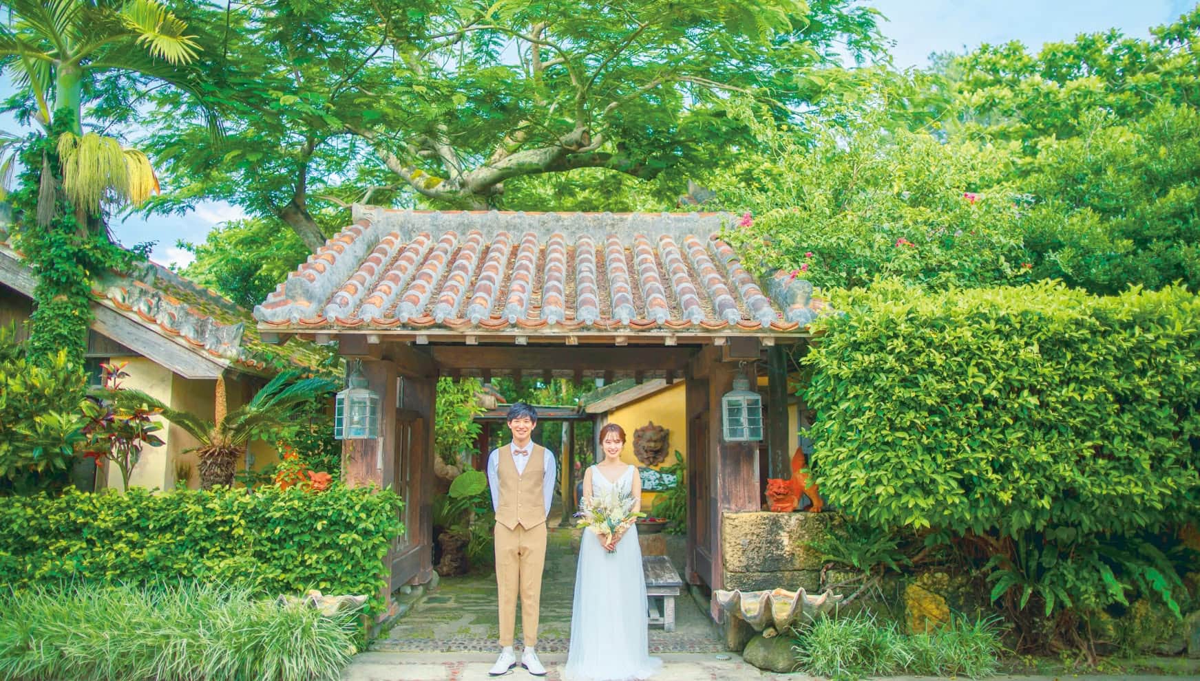 石垣島の「舟蔵の里」にある、趣ある赤瓦屋根の門の前に並んで立つ新郎新婦。豊かな亜熱帯の植物に囲まれ、シーサーが守る伝統的な琉球建築の風情と、ベージュのベスト姿の新郎、ナチュラルなウエディングドレス姿の新婦が調和した、石垣島らしい情緒あふれる一枚。