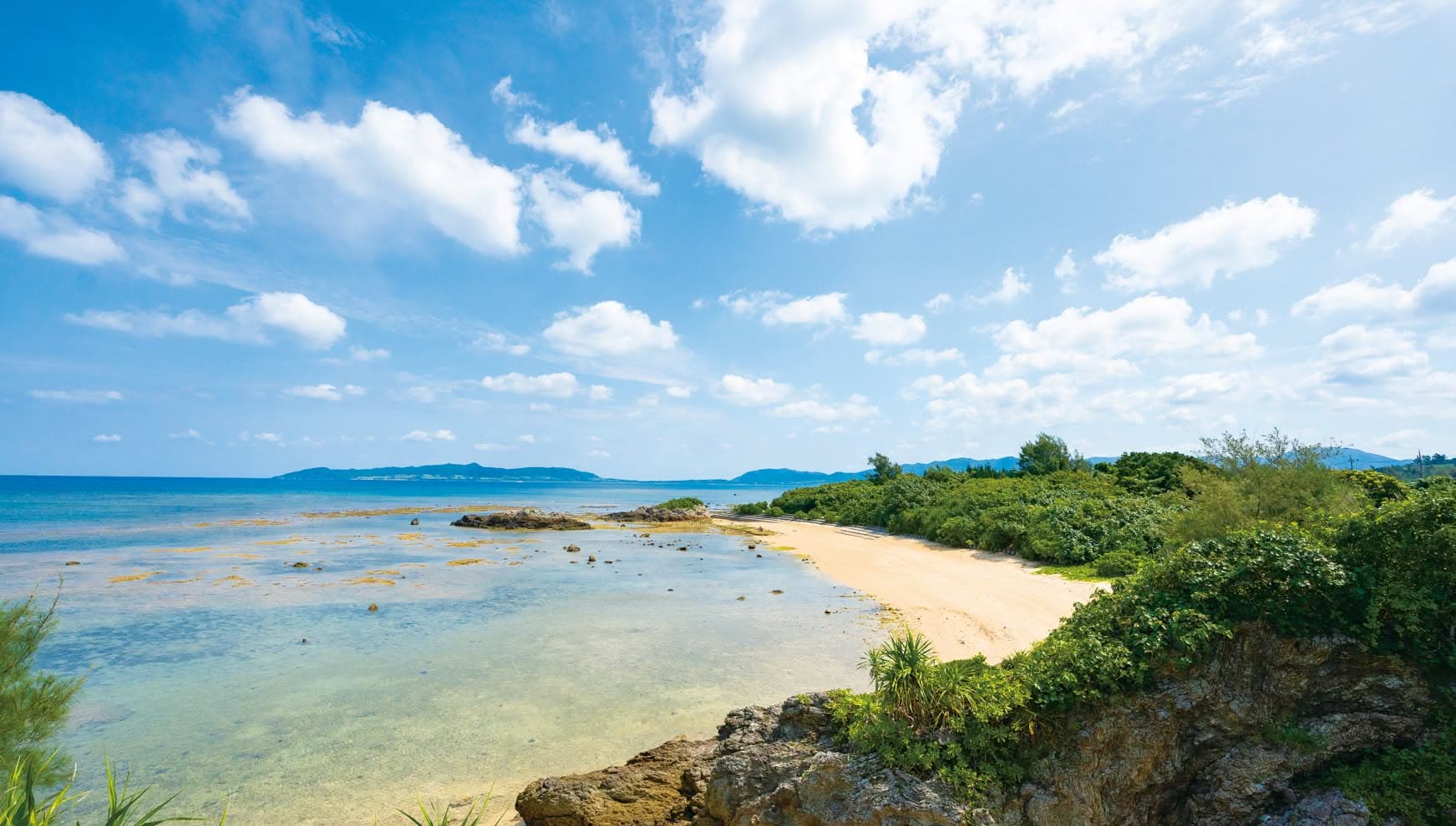 石垣島の「観音ビーチ」を望む高台からの絶景。透明度の高いエメラルドグリーンの浅瀬と、白い砂浜、そして海岸沿いに広がる力強い緑の植生が美しいコントラストを描く。遠くには島影が浮かび、大きな白い雲が浮かぶ青い空が開放感を引き立てるロケーション。