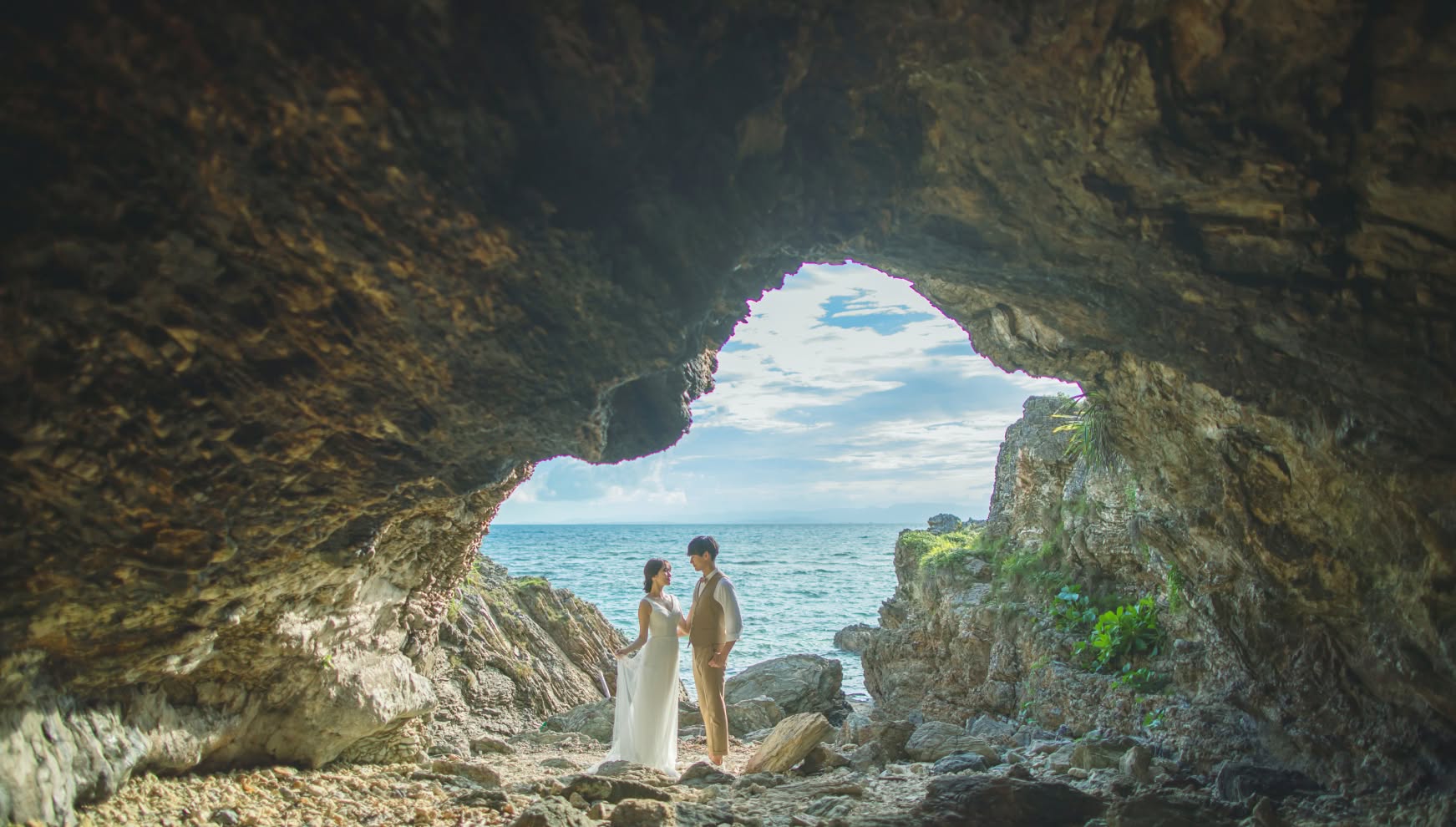 石垣島の「観音崎洞窟」内部から、天然の岩のフレーム越しに海を望む幻想的なウエディングフォト。洞窟のシルエットと外に広がる青い空、輝く海がコントラストを生み出し、その中心で見つめ合う新郎新婦がドラマチックに描かれた一枚。