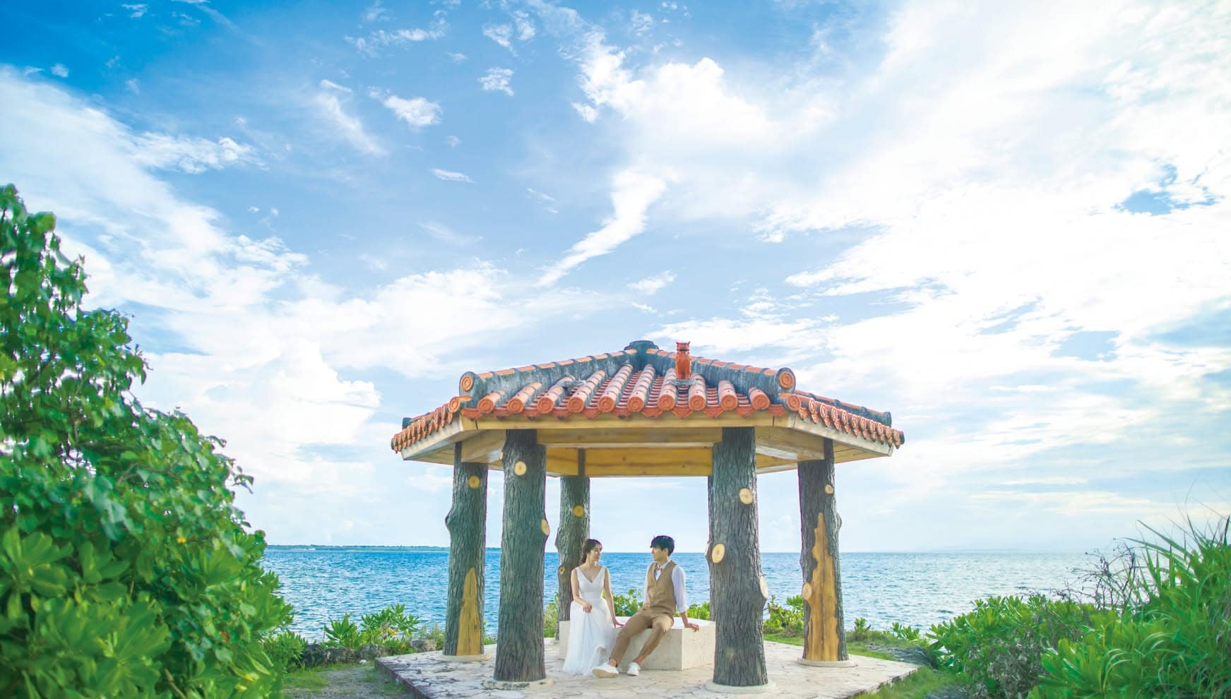 石垣島の「観音崎灯台」近くにある、赤瓦屋根の東屋（あずまや）で寄り添い座る新郎新婦。背景にはどこまでも続く青い海と空が広がり、沖縄らしい建築様式と自然が融合した、穏やかで心地よい雰囲気のウエディングフォト。