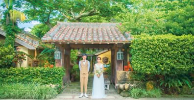 沖縄の伝統的な赤瓦の門を背景に、仲良く並んで立つ新郎新婦のウエディングフォト。豊かな緑に囲まれた古民家風のロケーションで、ベージュのベストを着用した新郎と、繊細なレースの白いドレスを纏った新婦が、温かな日差しの中で微笑む情緒溢れる一枚です。